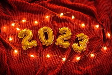 the numbers 2023 on the background of a red knitted cozy plaid and Christmas lights.