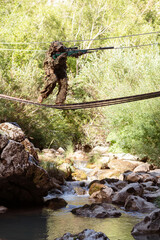 A military man or airsoft player in a camouflage suit sneaking the rope bridge and aims from a sniper rifle to the side or to target. 