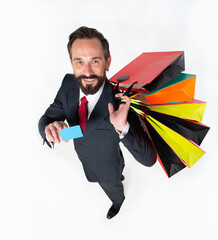 Top view of handsome bearded shopkeeper man smiling happy holding shopping bags and discount credit card. Attractive businessman in suit with shopping bags showing card in hand on white background. - obrazy, fototapety, plakaty