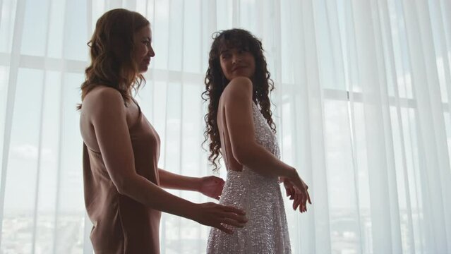 Happy mature Caucasian woman helping her daughter to zip sequin wedding dress then looking at her