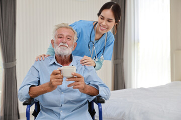Fototapeta premium Happy caregiver giving senior caucasian man with wheelchair a cup of coffee or tea for breakfast in bedroom at retirement house. Asian smiling nurse taking good help care and support elder patient.