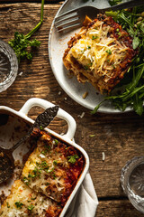 Baked Vegan lasagna casserole with roasted eggplant and mushrooms on a rustic wooden table served on a plate with some arugula  and a lot of vegan shredded cheese and fresh herbs on top