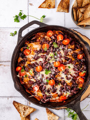 Mexican vegetarian dish with tortillas in an iron skillet on tiles, breakfast idea with black beans, cherry tomatoes, tortillas, and cheese, top view