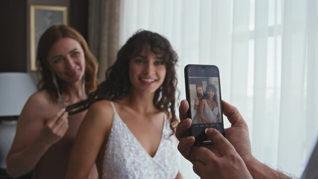 Unrecognizable man holding smartphone taking pictures of young bride and her mother, selective focus shot