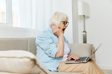 a shocked elderly woman is sitting on a comfortable sofa holding a laptop on her lap and looking very scared at the laptop monitor holding her hand near her face