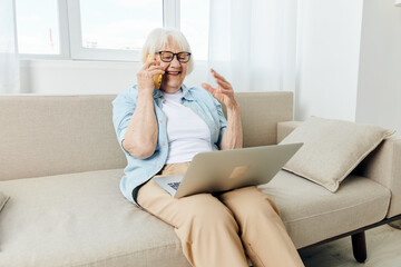 a cheerful, happy elderly woman is sitting on the couch working from home and talking on the phone with her hand raised, holding a laptop on her lap