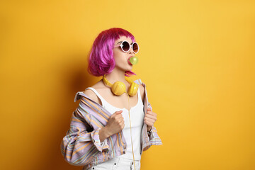 Fashionable young woman in colorful wig with headphones blowing bubblegum on yellow background,...