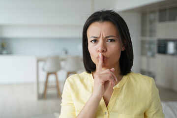 Fototapeta na wymiar Serious frowning young woman shows hush gesture, asking to keep privacy, secret and silence at home