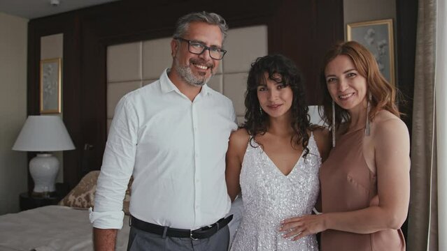 Medium family portrait of cheerful Caucasian bride and her parents standing indoors posing on camera