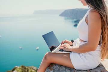 Naklejka premium Digital nomad, woman in the hat, a business woman with a laptop sits on the rocks by the sea during sunset, makes a business transaction online from a distance. Freelance, remote work on vacation.