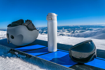 Ski helmet, thermos and goggle on snowboard on snowy mountain top
