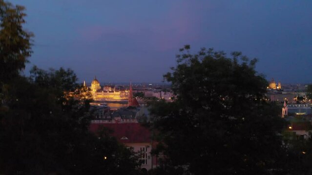 Drone taking off and showing Budapest  at night through the trees. The iluminated Parliament beyond the Danube River