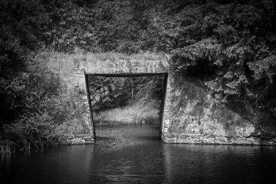 Grayscale shot of water flowing under a stone bridge with greenery around