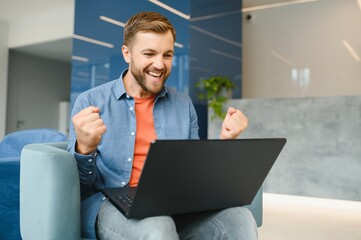 happy smiling remote online working man in casual outfit with laptop in joyful successful winning gesture sitting in an coworking office at a work desk.