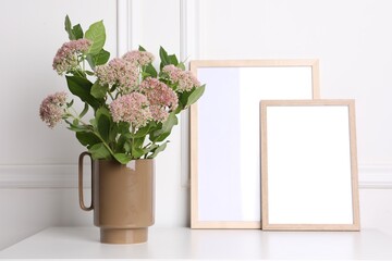 Stylish ceramic vase with beautiful flowers and blank frames on table near white wall