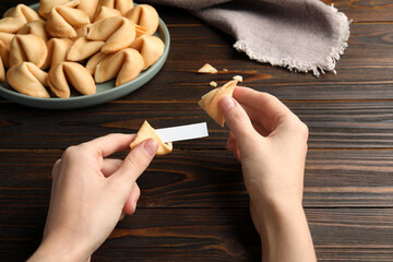 Woman holding tasty fortune cookie and paper with prediction at wooden table, closeup. Space for...