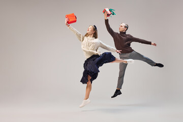Young and graceful couple of ballet dancers in warm winter clothes dancing with festive gift boxes on grey background. Christmas, New year, holidays, party concept