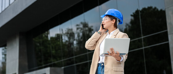 Female industrial engineer with laptop talking by mobile phone at construction site
