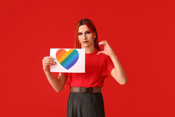 Stylish transgender woman holding paper sheet with rainbow heart against red background