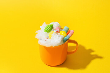 Cup with sweet cotton candy and different sweets on yellow background