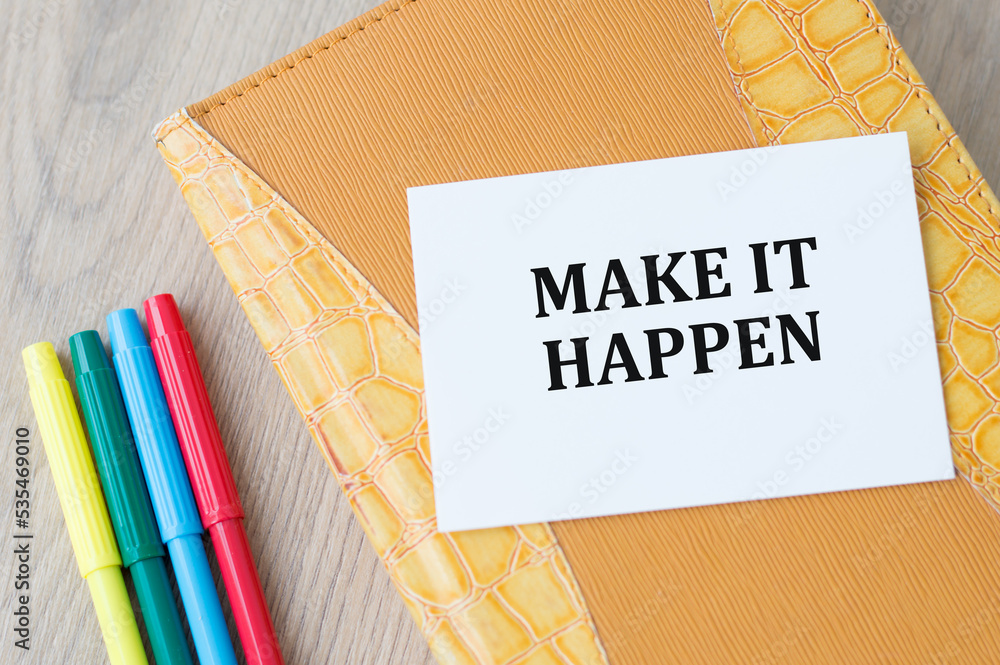Poster on the card the text make it happen on the background of a notepad on a wooden table next to colored