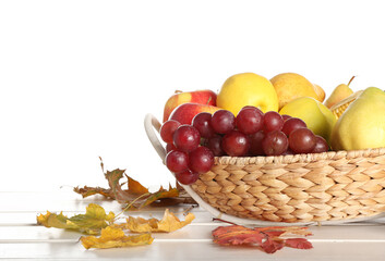 Wicker bowl with fresh fruits and autumn leaves on table against white background, closeup. Harvest festival