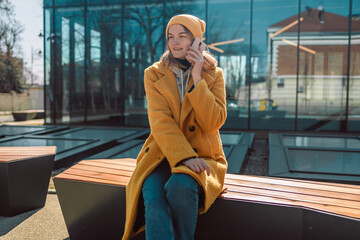 Fototapeta premium Young smiling caucasian fun woman in bright clothes sits on a stylish wooden bench in the city museum outdoors use mobile cell phone chat with boyfriend on social network. People active urban