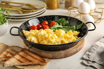 Wooden board with frying pan of scrambled eggs, tomatoes, greens and toasts on light table
