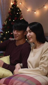 Vertical Screen: Korean couple watch tv holding remote control in hand on sofa at home in new year holidays. side view husband chatting with his wife discussing about talk show and laughing