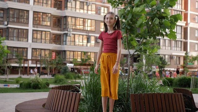 A preteen girl is dancing and fooling around standing on a bench in the yard against the background of apartment buildings and a playground.
