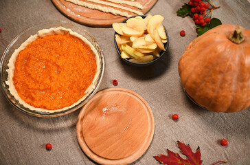 Top view festive Thanksgiving pie, pastry stripes on wooden board, slices of apple in bowl, autumn maple leaves, viburnum berries, whole ripe pumpkin on a linen tablecloth. Rustic still life with food