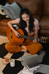 Cheerful woman learning to play guitar, using laptop computer on carpet in cozy living room