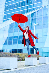 Blue Monday concept. Young executive business woman playing with a red umbrella on a rainy day. She wears a casual urban look, a red coat, jeans and a white blouse.