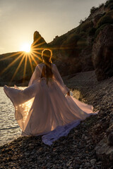Fototapeta na wymiar A mysterious female silhouette with long braids stands on the sea beach with mountain views, Sunset rays shine on a woman. Throws up a long white dress, a divine sunset.