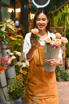 Positive young woman small business entrepreneur wearing apron holding bouquet of pink roses standing in front of flower shop