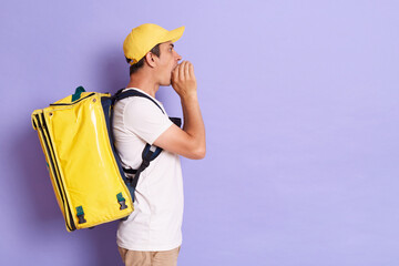 Side view of delivery employee man in cap, holding thermal bag backpack with food, working as courier, posing isolated on violet background, keeps hands near mouth, screaming, announcing.