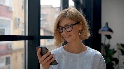 Woman holding smartphone, reading a message. Attractive blonde manager portrait on background of windows. Office room and worker with mobile phone. Concept of business and technology 