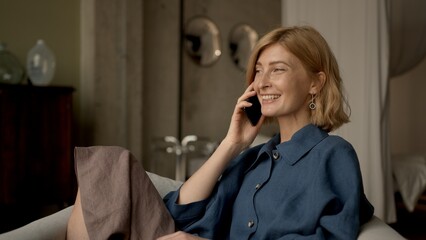 Blonde woman talk on the phone sitting in armchair, portrait of happy woman making a call. Gimbal shot of a cheerful and smiling adult person using phone. 