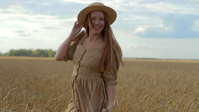 Portrait of an attractive young woman in a summer dress and hat walking in wheat field turns around against background of beautiful sunset looks into camera and laughs. Happy person outside