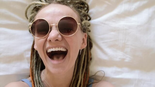 Happy smiling girl joyfully falling on comfy bed in hotel room in summer vacation, young crazy woman with dreadlocks taking selfies in round sun glasses, making excited selfie video call, slow motion