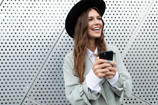 portrait of a strong girl in an autumn coat and a black hat against the background of an aluminum wall of a building with a mobile phone in her hands, Concept freelancing and remote work