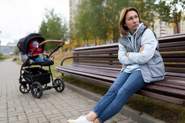 Fototapeta na wymiar upset mother sits on a bench separated from the pram with her baby, the concept of postpartum depression