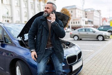 adult gray-haired man speaks on a mobile phone at the open hood of a broken car, the concept of life and vehicle insurance against accidents