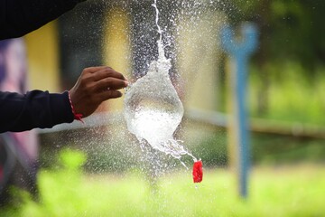 Person popping a red balloon filled with water with a blurry park in the background.