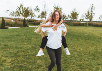 A happy mother and a little girl play and have fun in the park together, a girl rides on her mother's back. people in white t-shirts