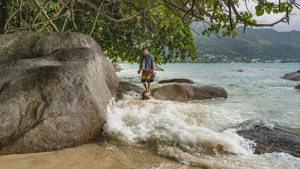 A man stands on a boulder in the surf zone of the ocean and looks in surprise at the foam of waves crashing on the rocks. A towel is on his shoulders, and he holds flip-flops in his hands. Seychelles.