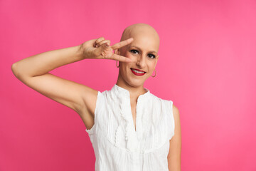 a woman suffering from cancer with a shaved head and a victory gesture on a pink background