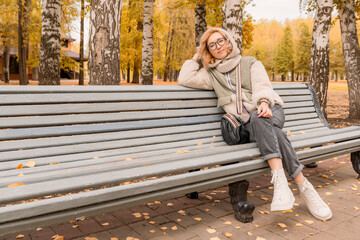 Fototapeta premium Young and beautiful blonde girl in glasses and insulated clothes sits on bench in autumn recreation park against background of yellowing trees. Looks at camera.