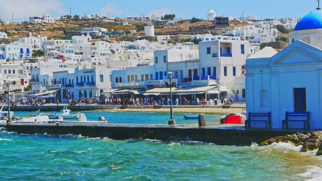 Mykonos port panorama with Greek Orthodox church. Traditional white buildings on embankment and boats on quay. People walking on bay, famous Cyclades Island