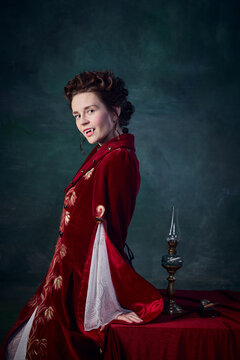 Portrait of beautiful woman with white pale skin in image of medieval vampire posing in elegant red dress over dark green background